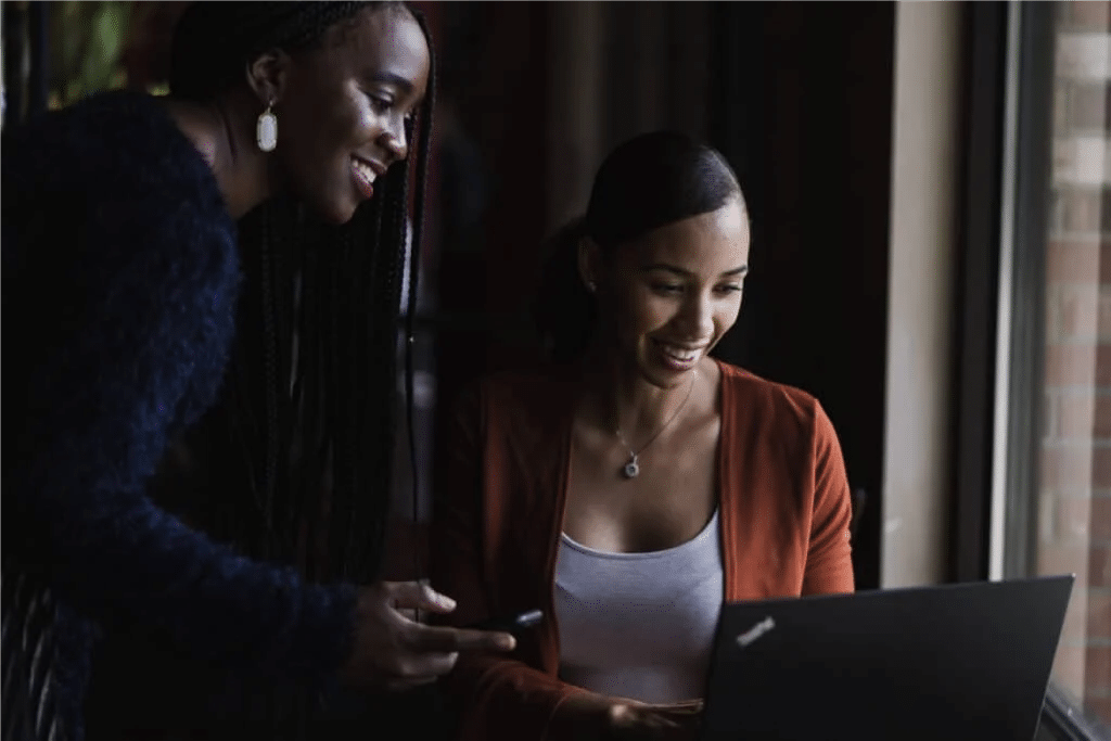 Two women smiling while using a laptop by window